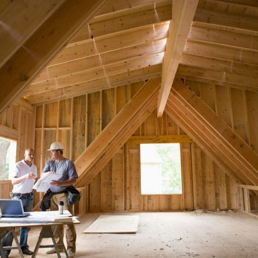 Two men review blueprints in wooden attic space.