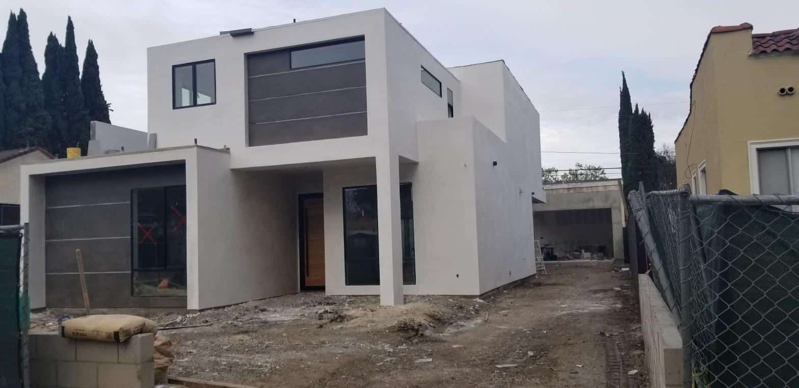 Two-story modern white stucco home under construction in Los Angeles with bare dirt yard and chain-link fence