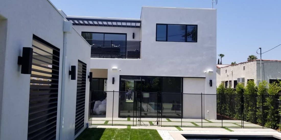 Modern two-story white stucco house with large sliding glass doors, second-floor balcony, pool fence, and manicured lawn.