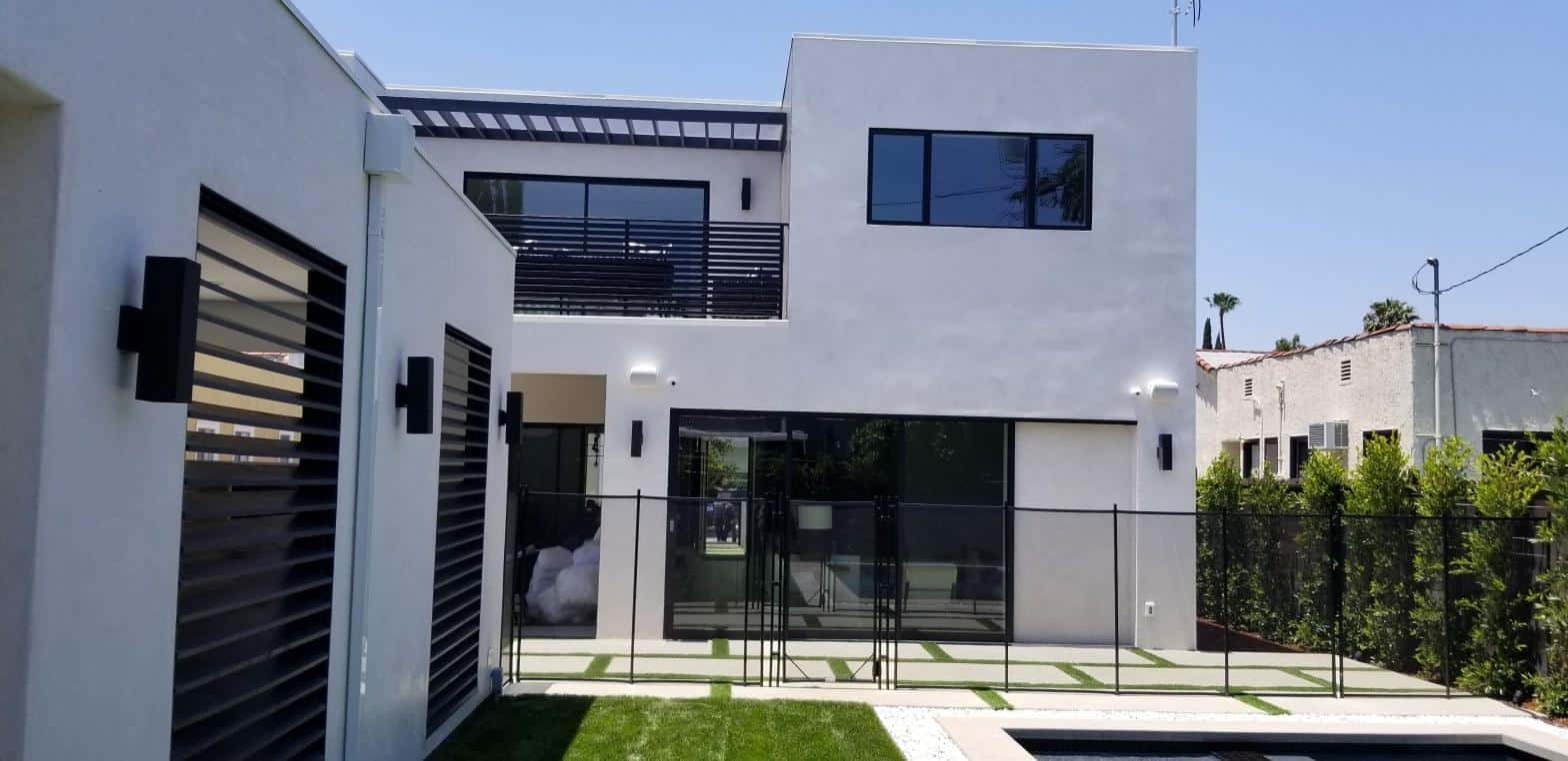 Modern two-story white stucco house with large sliding glass doors, second-floor balcony, pool fence, and manicured lawn.