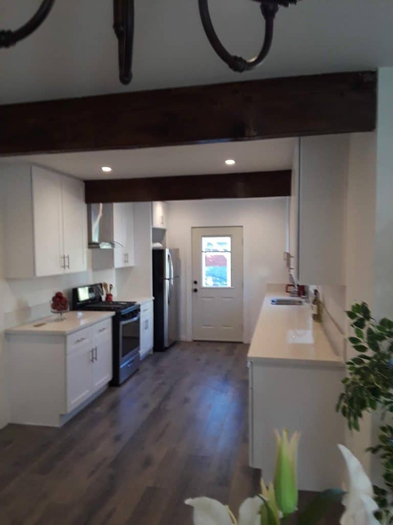Remodeled galley kitchen featuring white shaker cabinets, gas range, stainless refrigerator, dark wood ceiling beams, and hardwood floors.